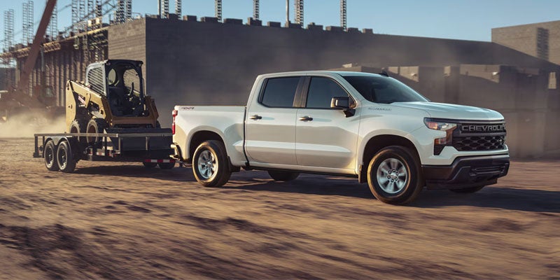 A silver 2026 Chevrolet Silverado tows a yellow excavator behind it on a construction site on a sunny day in City of Industry, CA