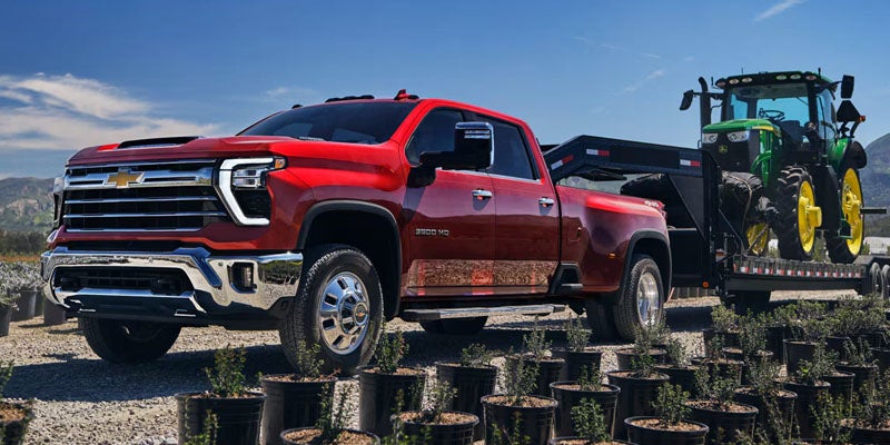 Red Chevrolet Silverado HD pickup truck towing a trailer with a tractor at a construction site.